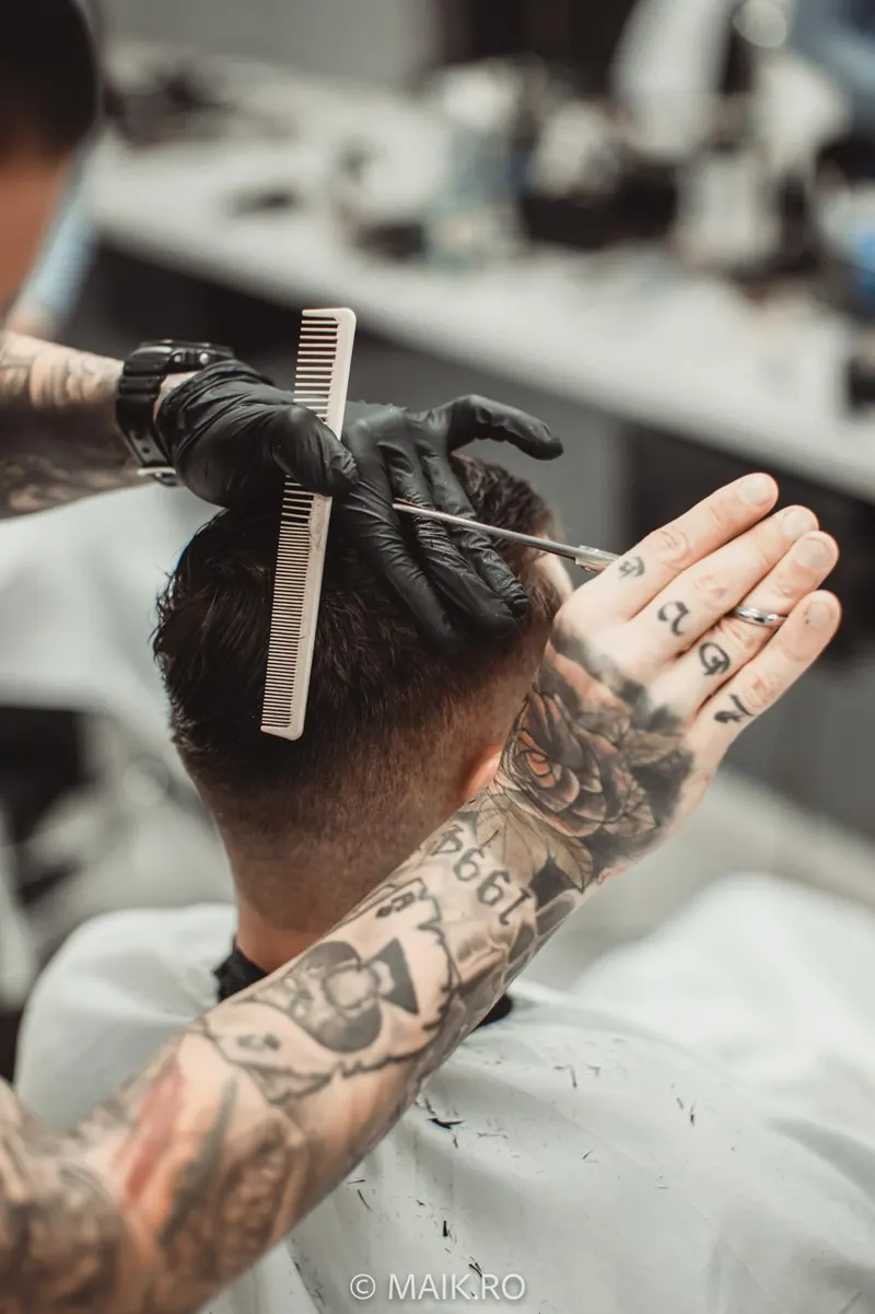 Mâinile tatuate ale frizerului tăind cu foarfeca peste pieptene, la ceafa unui client.