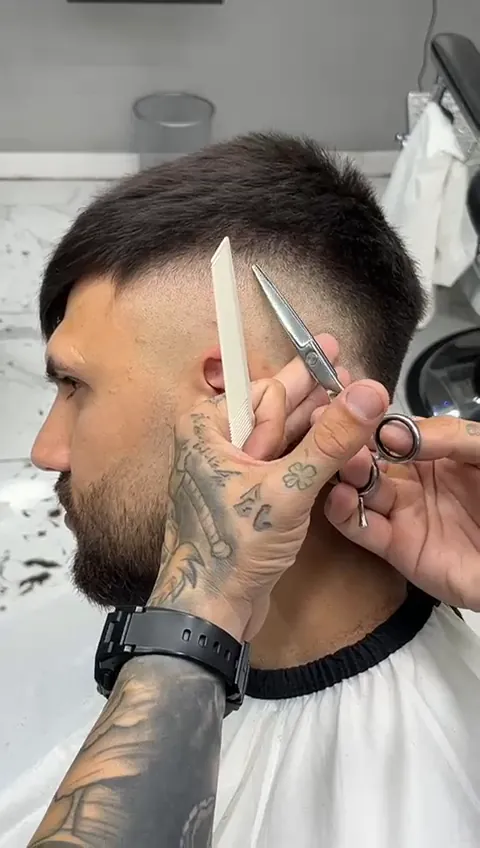 Frizerul tunde cu foarfeca peste pieptene, deasupra urechii unui client cu barbă; laterale în fade.